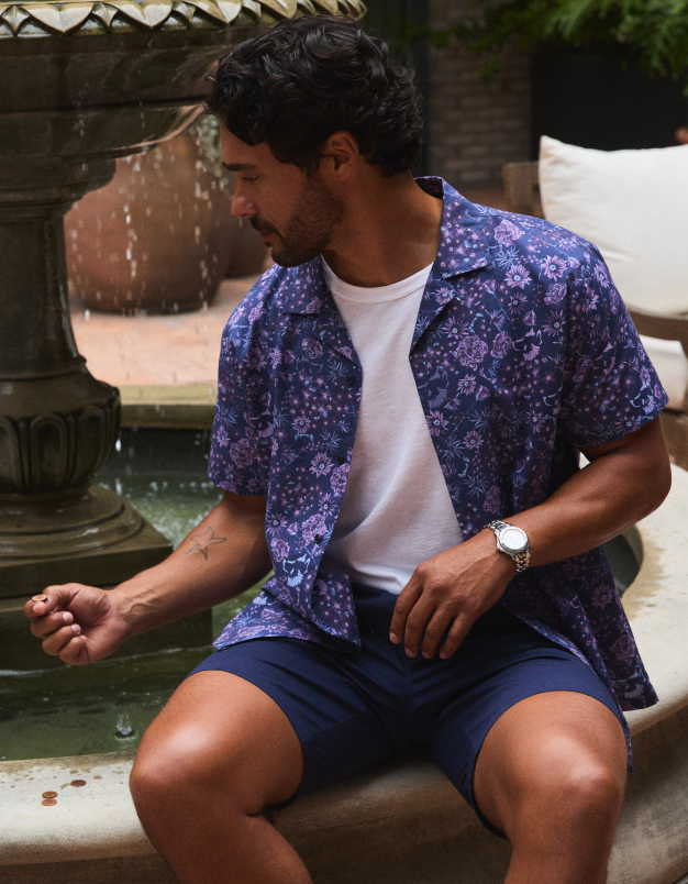 Man wearing a blue floral shirt and navy shorts sitting by a fountain.